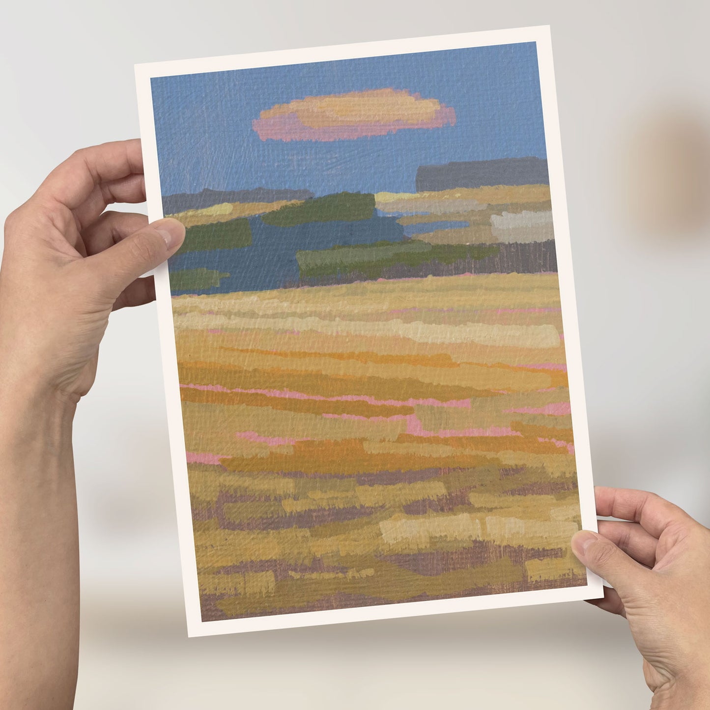 Hand hält Mini-Poster mit malerischer Herbstfeld-Landschaft und Wolkenhimmel im A4 Format.